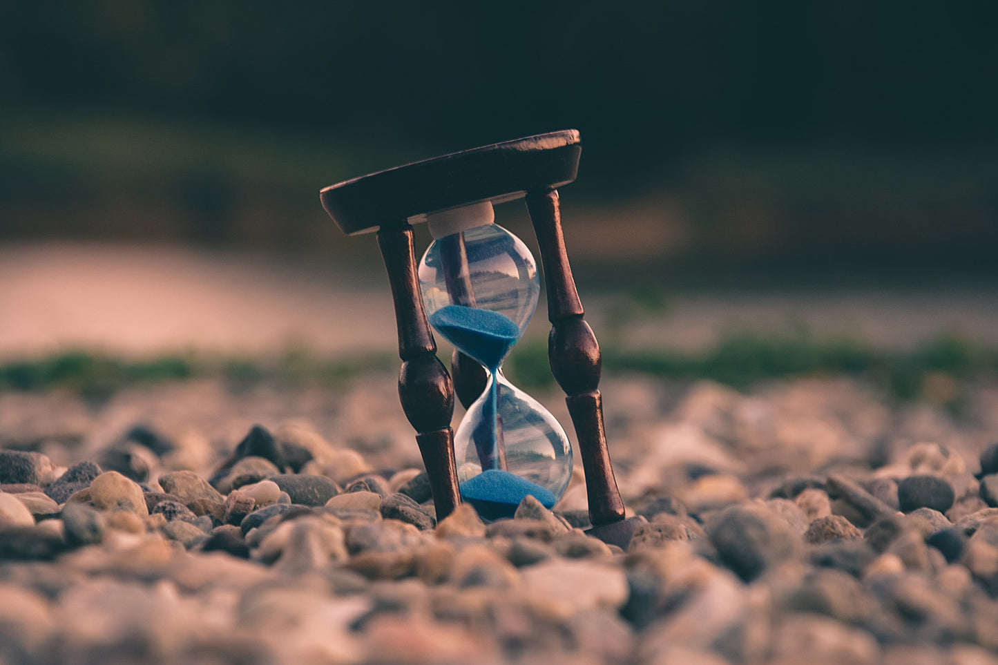 An hourglass full of blue sand sits tilted in a patch of pebbles.