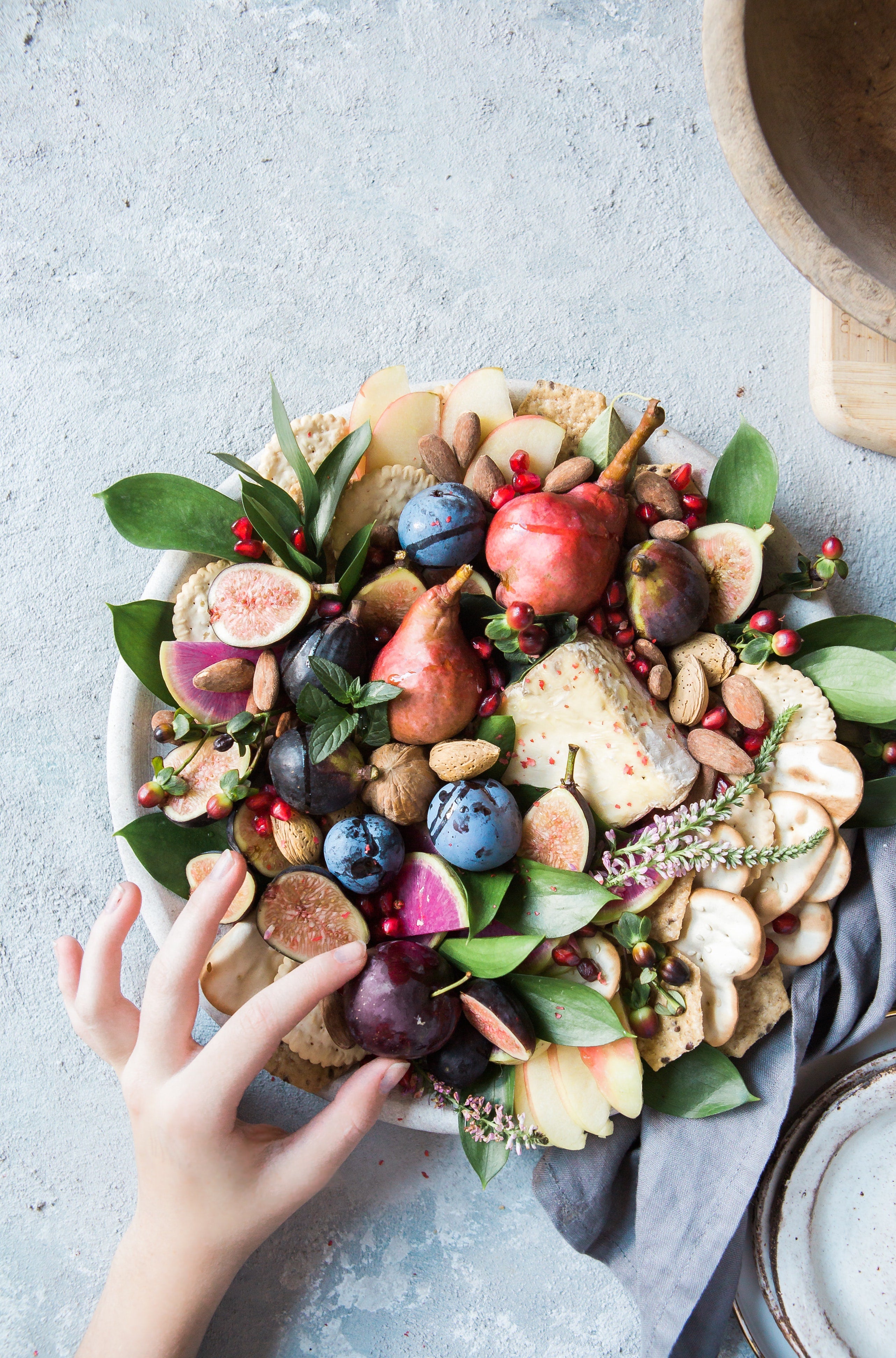 A hand carefully plucks a plum from a spread of fruits, nuts, and cheeses. 