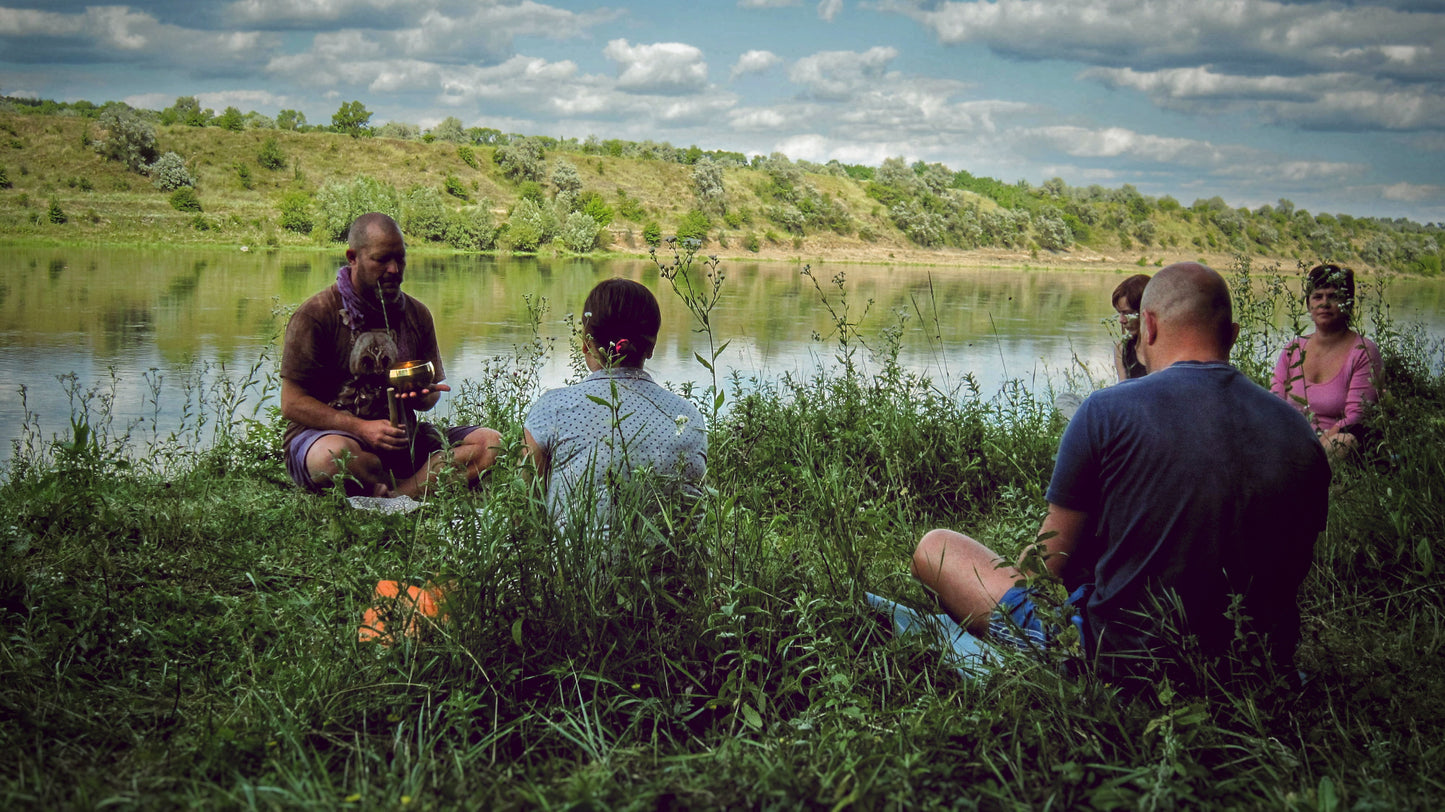 A small group of people sit in tall grass alongside a riverbank as a sound healer shows them a singing bowl.